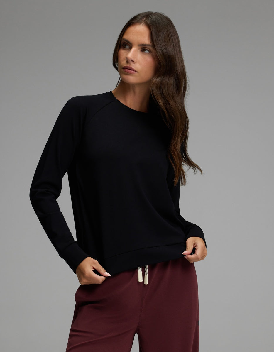 A woman with long brown hair wears the Cozy Earth Women’s Brushed Bamboo Pullover Crew in black and maroon drawstring pants, standing against a plain grey background and looking to her right with a neutral expression.