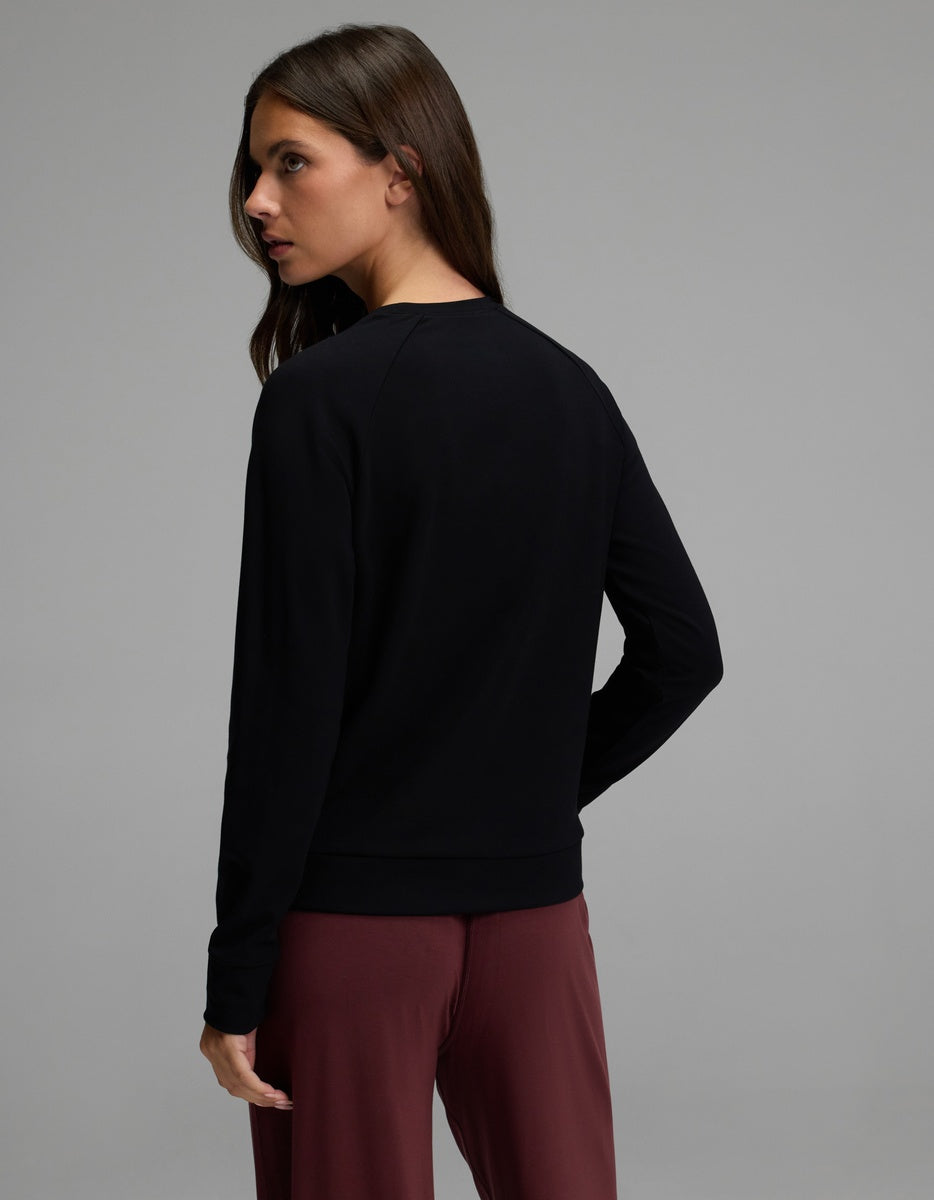 A woman with long brown hair wears the Cozy Earth Women’s Brushed Bamboo Pullover Crew in black, paired with maroon pants. She stands against a plain gray background, looking over her left shoulder with her back mostly facing the camera.