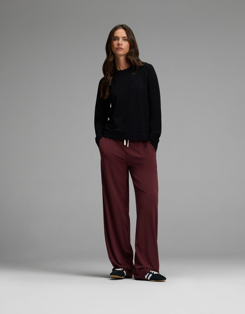 A woman stands with her hands in her pockets, wearing the Cozy Earth Women’s Brushed Bamboo Pullover Crew in black, loose burgundy pants, and black Adidas slides against a plain gray background.