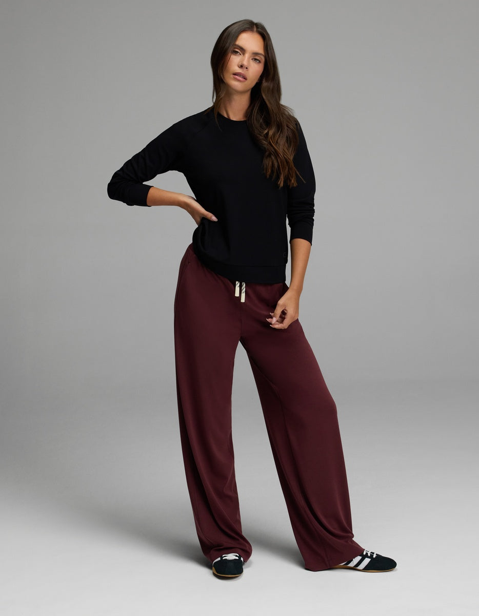 A woman stands in a studio wearing the Cozy Earth Women’s Brushed Bamboo Pullover Crew, paired with loose maroon drawstring pants and black-and-white sneakers. She has long brown hair and a neutral expression.