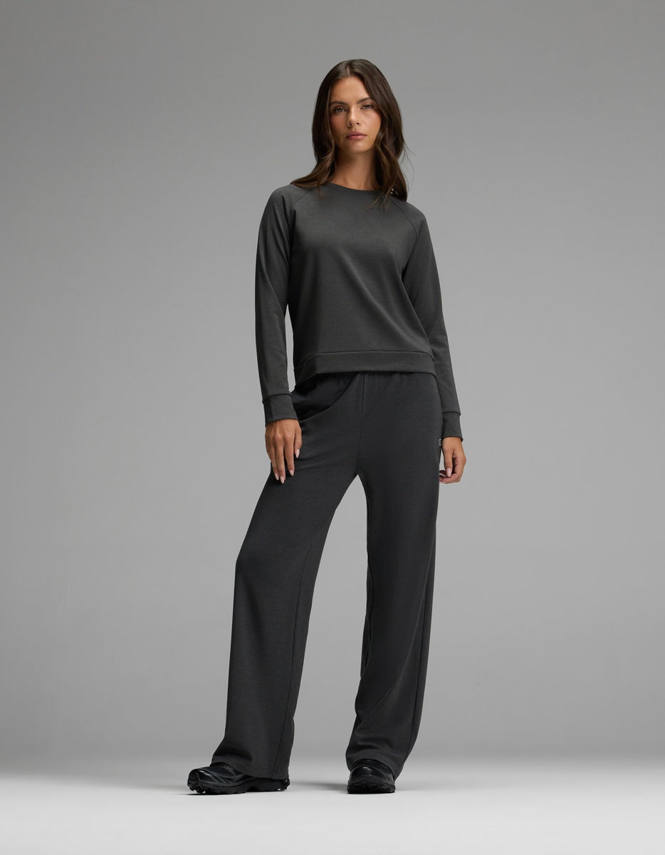 A woman stands against a gray background wearing the Cozy Earth Women’s Brushed Bamboo Pullover Crew in dark gray, paired with matching wide-leg pants, her posture relaxed and expression neutral.