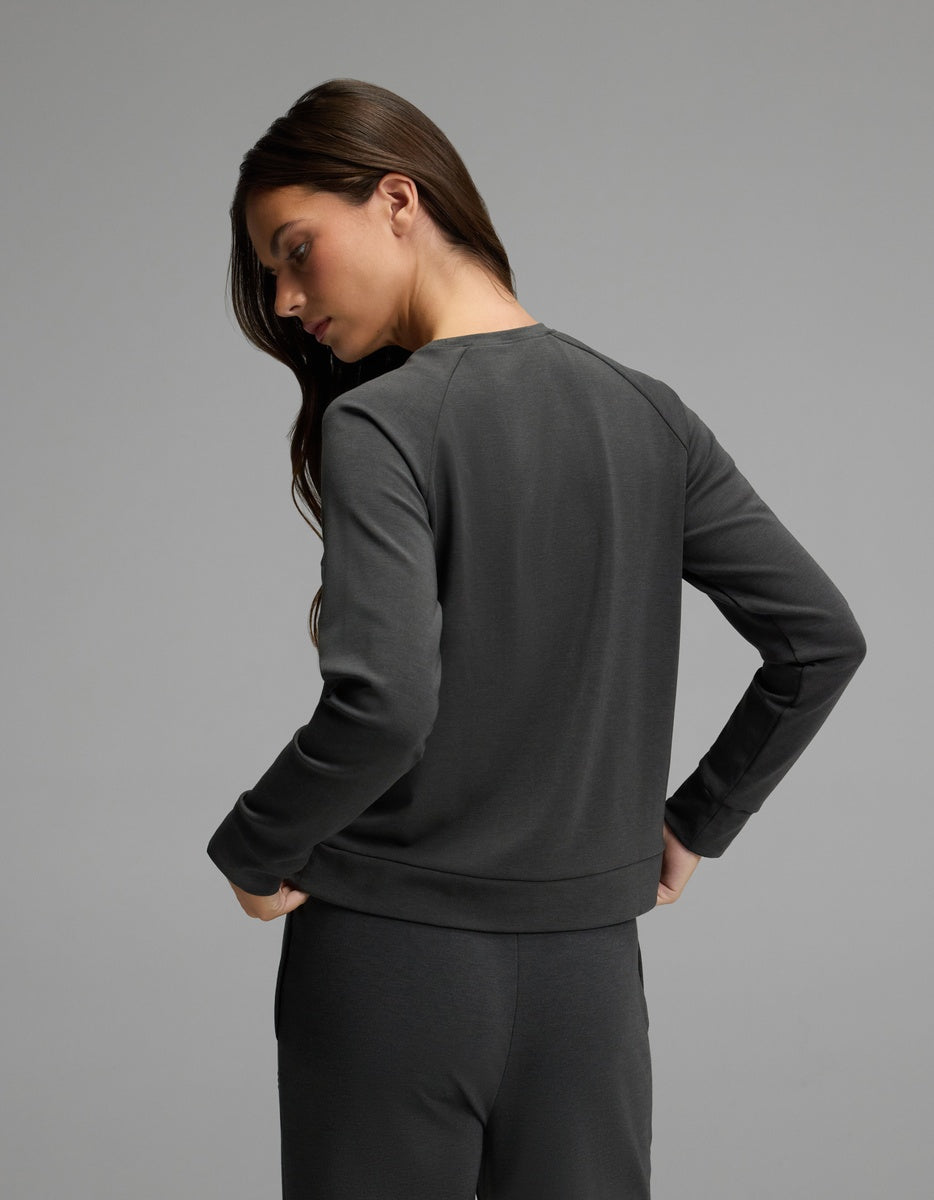 A woman with long brown hair, wearing the Cozy Earth Women’s Brushed Bamboo Pullover Crew in dark gray, stands facing away and looks over her shoulder against a plain gray background.
