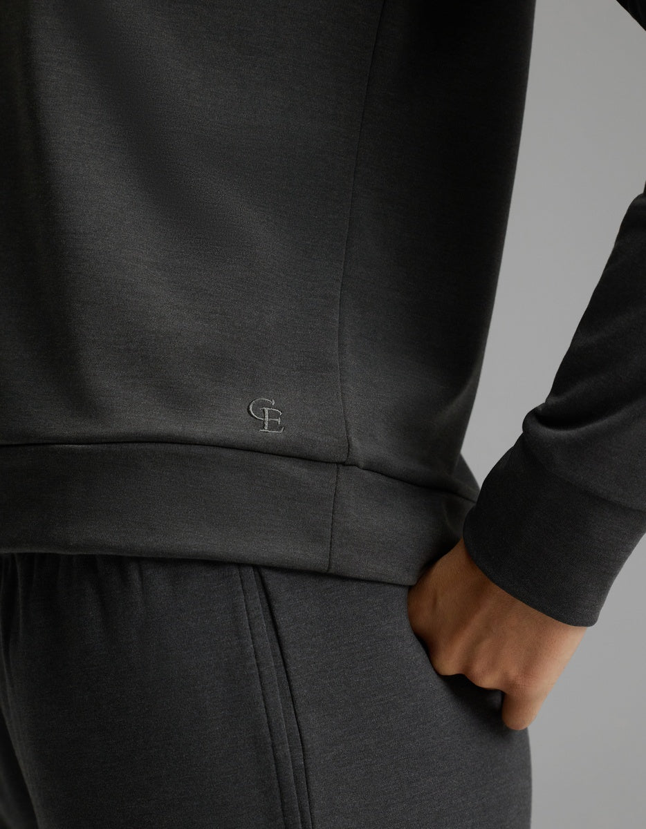 A person is wearing the Cozy Earth Women’s Brushed Bamboo Pullover Crew and matching pants, with one hand in the pocket. The dark gray sweatshirt features small “CE” initials stitched near the hem, set against a plain gray background.