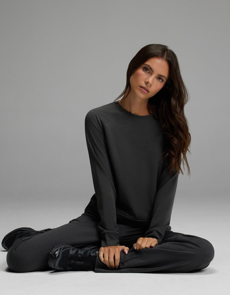 A woman with long brown hair sits cross-legged on the floor, wearing the Cozy Earth Women’s Brushed Bamboo Pullover Crew and matching pants, looking slightly to the side against a plain gray background.
