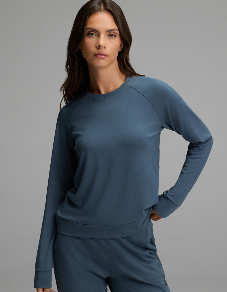 A woman with long brown hair wears the Cozy Earth Women’s Brushed Bamboo Pullover Crew in blue, standing against a plain grey background with a neutral expression.|Color:Dawn