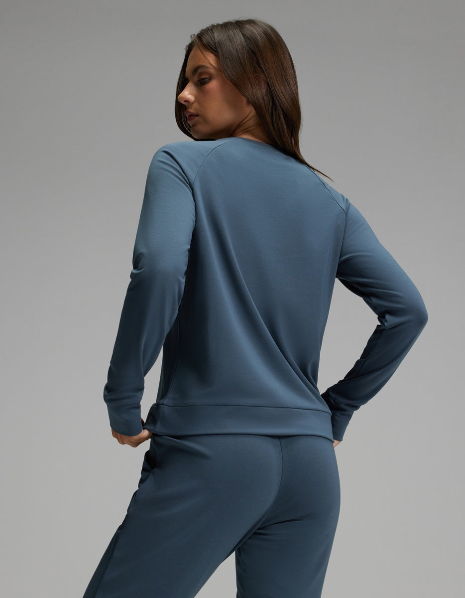 A person with long brown hair is wearing the Cozy Earth Women’s Brushed Bamboo Pullover Crew in blue, paired with matching pants, and is standing with their back turned while looking over their shoulder against a plain gray background.