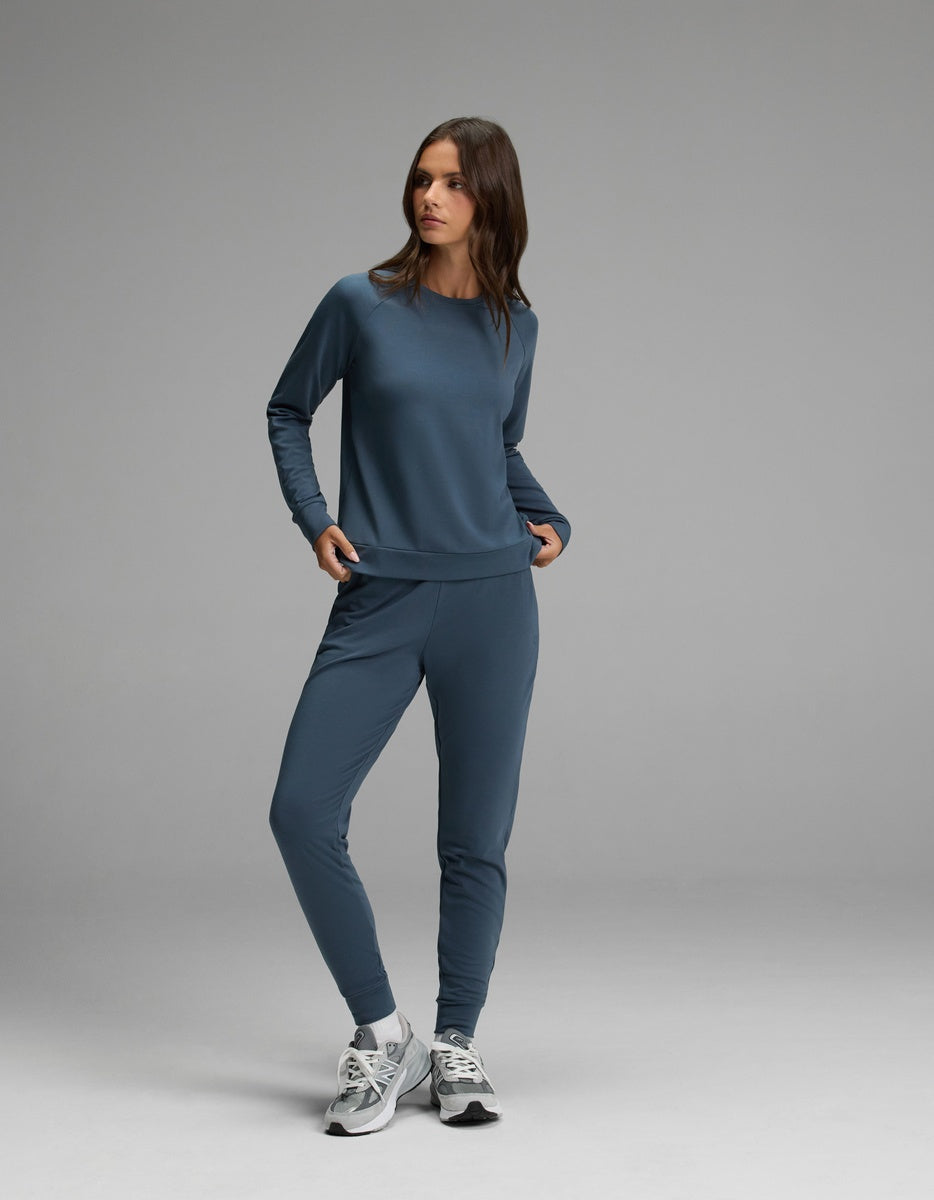A woman wearing the Cozy Earth Women’s Brushed Bamboo Pullover Crew in blue with matching joggers and gray sneakers stands against a plain gray background, looking slightly to her left.