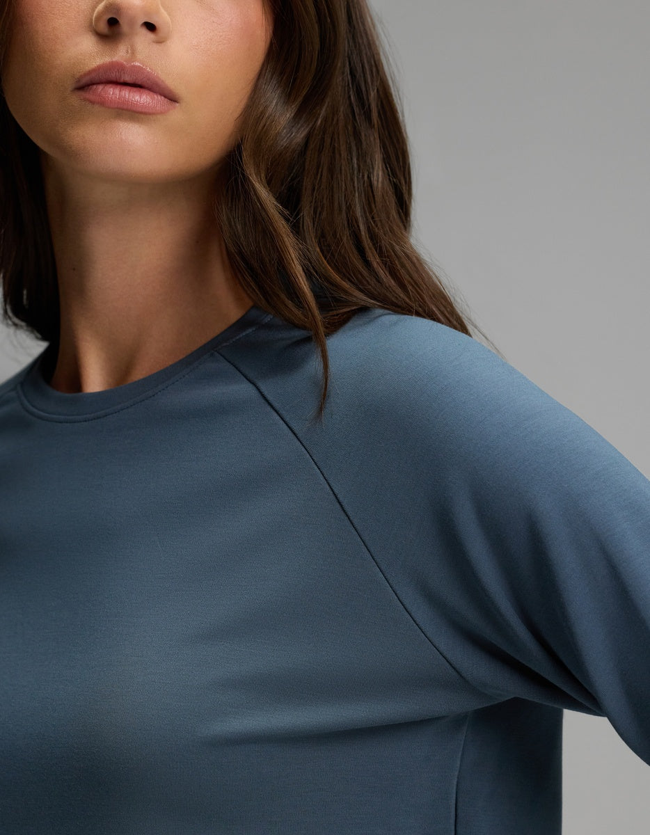 A woman with long brown hair is shown from the shoulders up against a plain gray background, wearing the Cozy Earth Women’s Brushed Bamboo Pullover Crew in blue.
