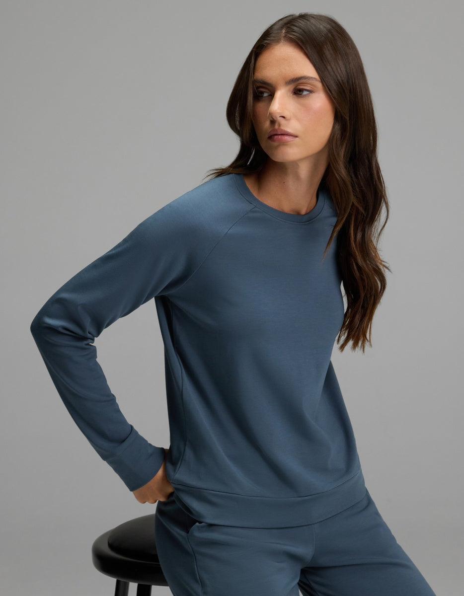 A woman with long brown hair, dressed in the Cozy Earth Women’s Brushed Bamboo Pullover Crew in blue and matching pants, sits sideways on a black stool, looking to her left against a plain gray background.