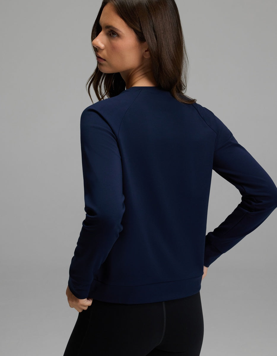 A woman with long brown hair, wearing the Cozy Earth Women’s Brushed Bamboo Pullover Crew in navy and black pants, stands facing away and looks over her left shoulder against a neutral gray background.