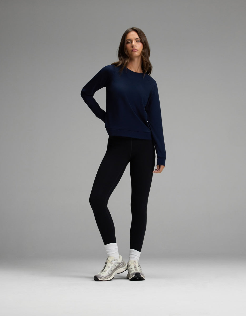 A woman stands against a gray background wearing the Cozy Earth Women’s Brushed Bamboo Pullover Crew, styled with black leggings, white socks, and white sneakers. She poses with one hand on her hip and a relaxed, confident expression.