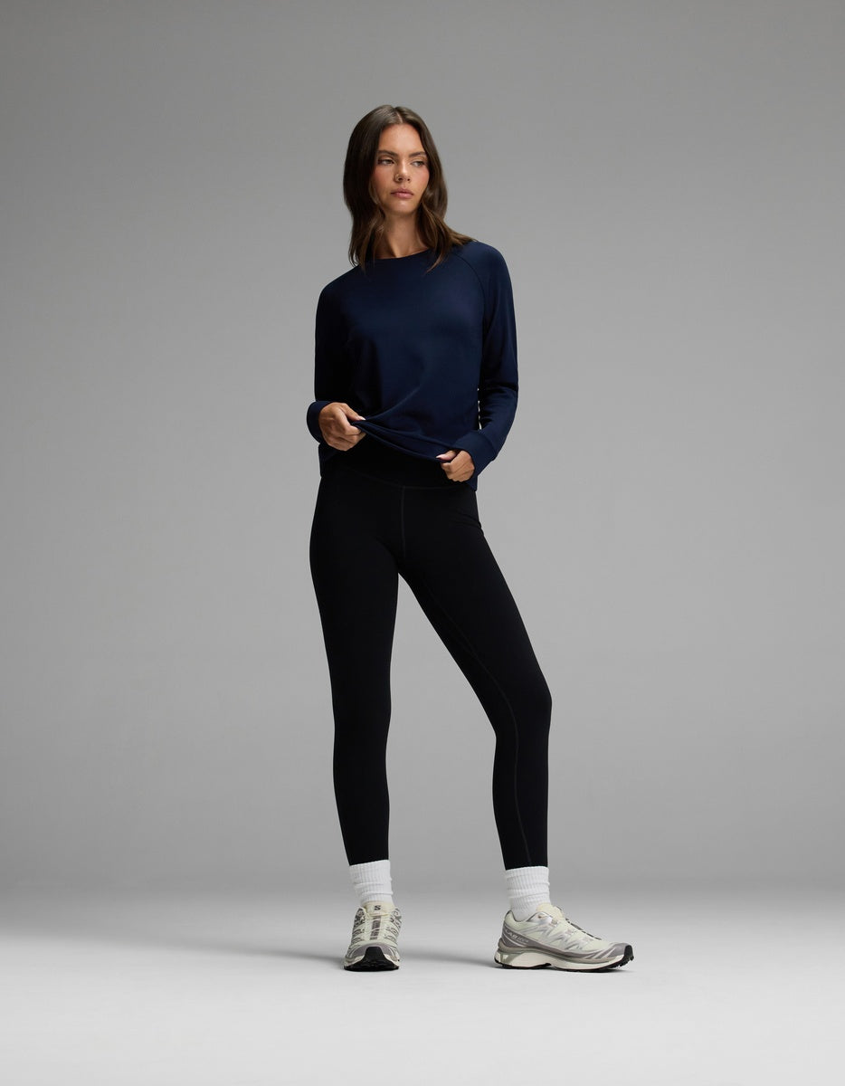 A woman stands against a plain gray background, wearing the Cozy Earth Women’s Brushed Bamboo Pullover Crew in navy, paired with black leggings, white socks, and white sneakers. She holds the hem of her pullover and looks slightly to the side with a neutral expression.