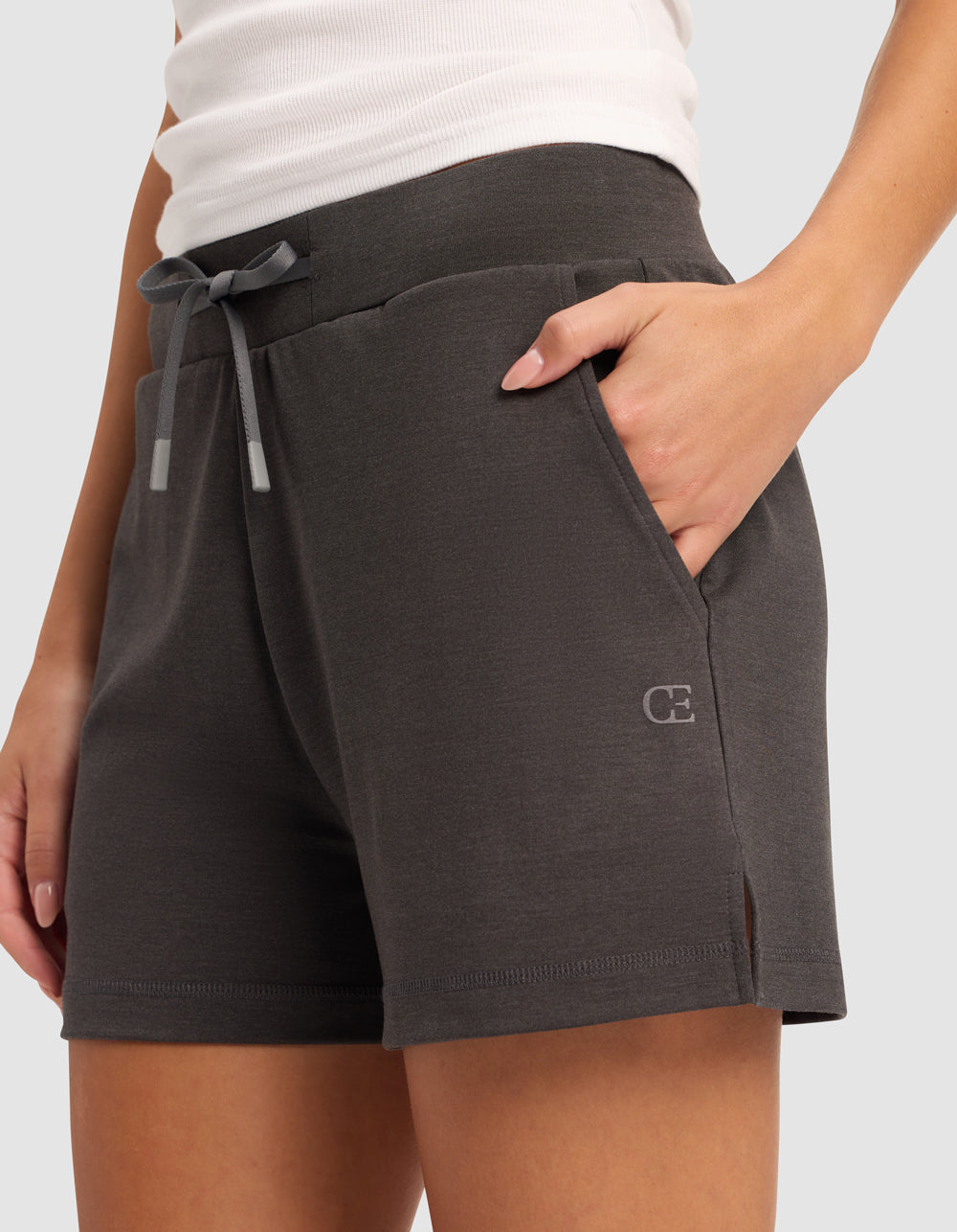A person wears Cozy Earth Women’s Brushed Bamboo Shorts in dark gray, featuring a small left-leg logo and pockets, paired with a white tank top. Their left hand is tucked into the shorts’ pocket.|Color:Charcoal