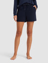A barefoot person wears Cozy Earth Women’s Brushed Bamboo Shorts with a long-sleeve navy top, hands in pockets, against a plain light background; the person's head is not visible.|Color:Navy