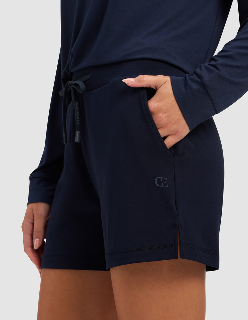 A woman wears Cozy Earth's Women’s Brushed Bamboo Shorts in navy blue, paired with a matching long-sleeve top and one hand in her shorts pocket, standing against a plain light background.