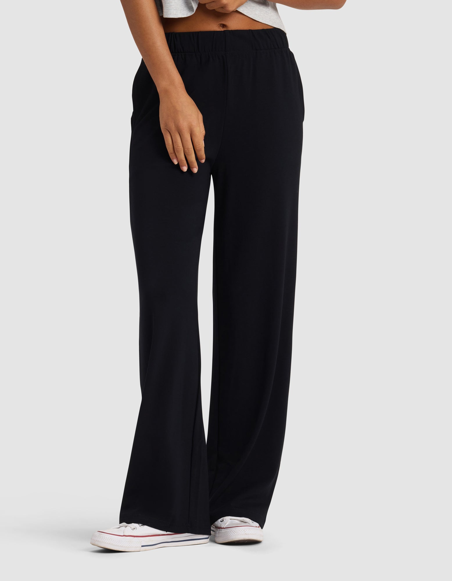 A person wears Cozy Earth's Women's Brushed Bamboo Wide Leg Pull-On Pant in black, paired with a light gray crop top and white sneakers, standing against a plain light background. Only the lower torso and legs are visible.|Color:Black