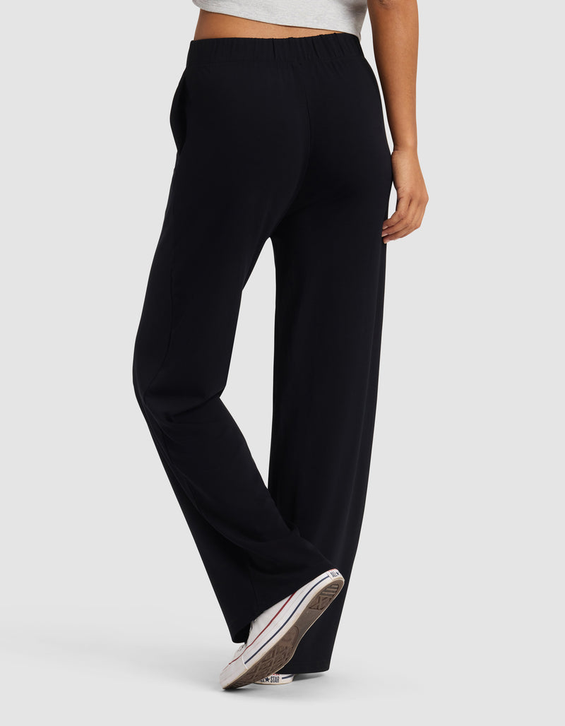 A person is seen from behind against a light gray background wearing Cozy Earth's Women's Brushed Bamboo Wide Leg Pull-On Pant in black, paired with a gray cropped top and white sneakers.