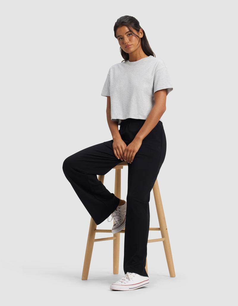 A woman in black Cozy Earth Women's Brushed Bamboo Wide Leg Pull-On Pants, a light gray cropped t-shirt, and white sneakers sits on a stool against a plain background, gazing at the camera with a relaxed expression.