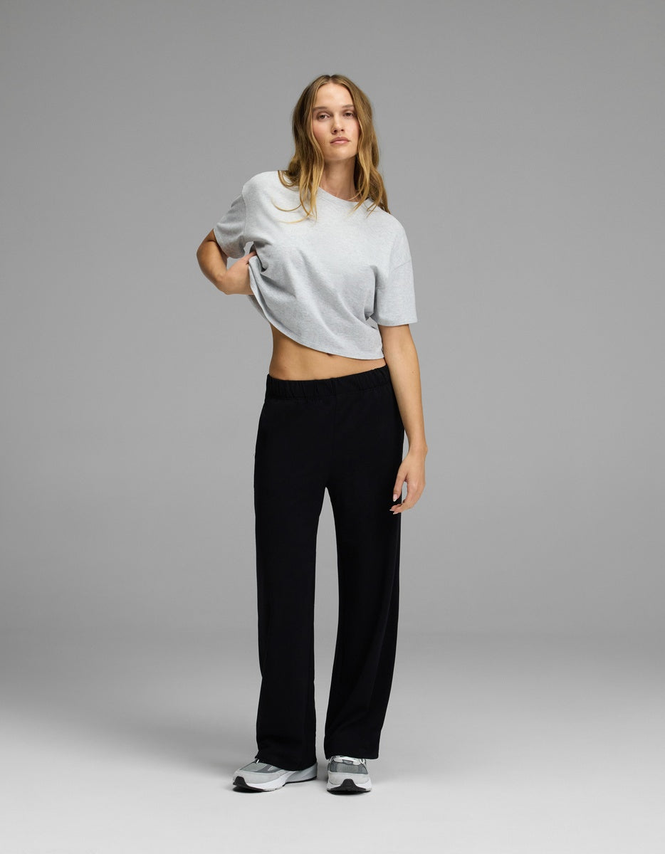 A person stands against a plain gray background, wearing a light gray t-shirt, gray sneakers, and Cozy Earth’s Women’s Brushed Bamboo Wide Leg Pull-On Pant in black. One hand rests on their hip while the other arm hangs loosely by their side.