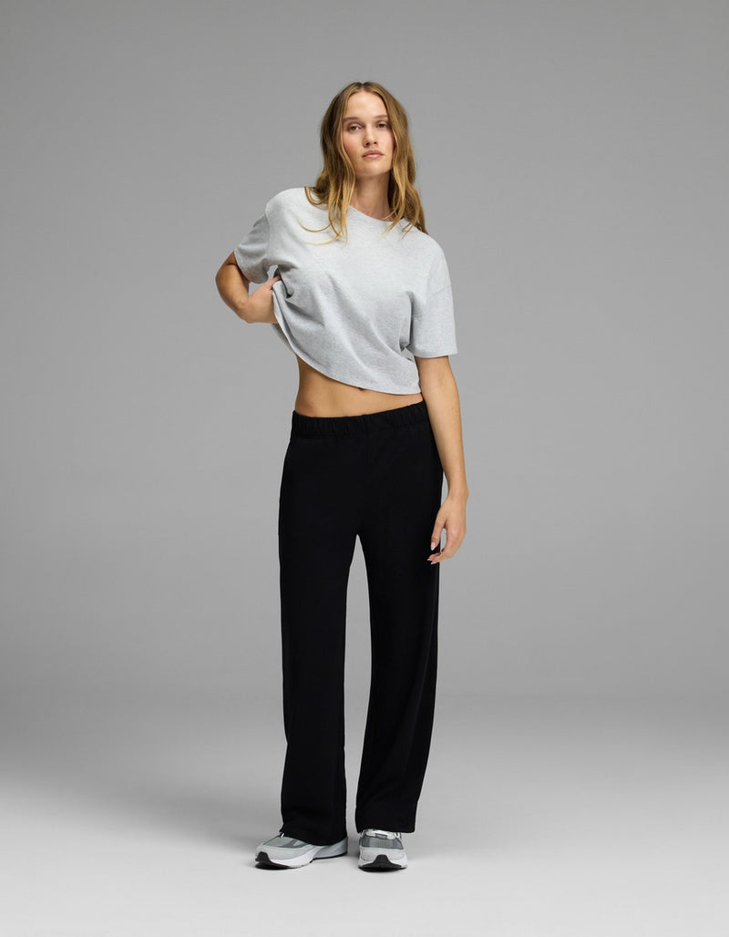 A person stands against a plain gray background, wearing a light gray t-shirt, gray sneakers, and Cozy Earth’s Women’s Brushed Bamboo Wide Leg Pull-On Pant in black. One hand rests on their hip while the other arm hangs loosely by their side.