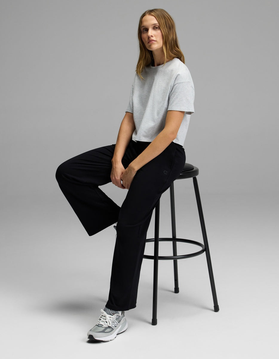 A person with long hair sits on a tall black stool, wearing a light gray T-shirt, Cozy Earth Women’s Brushed Bamboo Wide Leg Pull-On Pant in black, and gray sneakers, against a plain gray background.