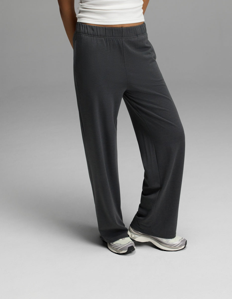 A woman wears Cozy Earth’s Women’s Brushed Bamboo Wide Leg Pull-On Pant in Charcoal, paired with white sneakers and a white top, standing with one leg crossed in front of the other against a plain gray background.