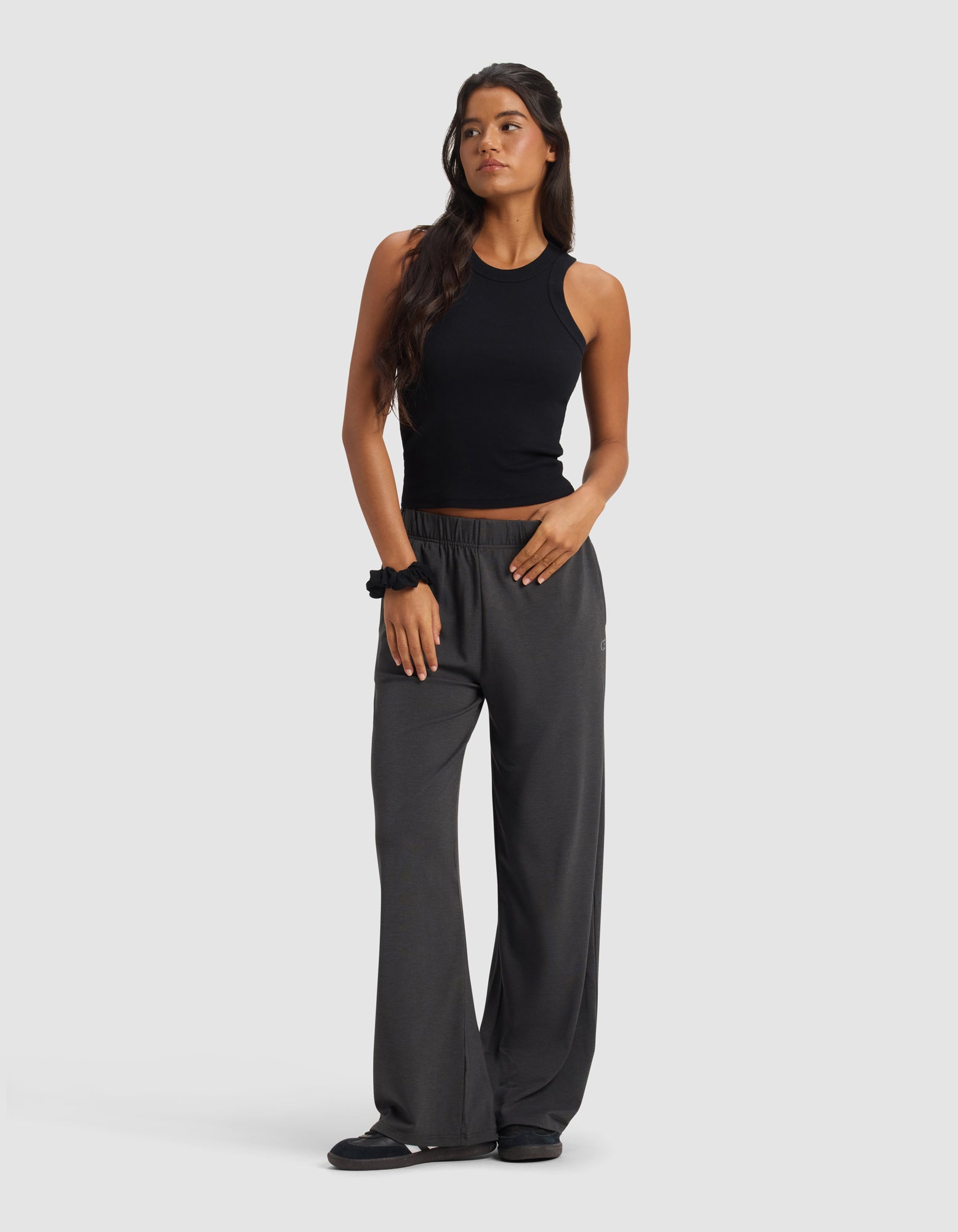 A woman with long dark hair wears Cozy Earth's Women's Brushed Bamboo Wide Leg Pull-On Pant in dark gray, paired with a black sleeveless top and black shoes. She poses against a light background, looking to the side in a relaxed manner.