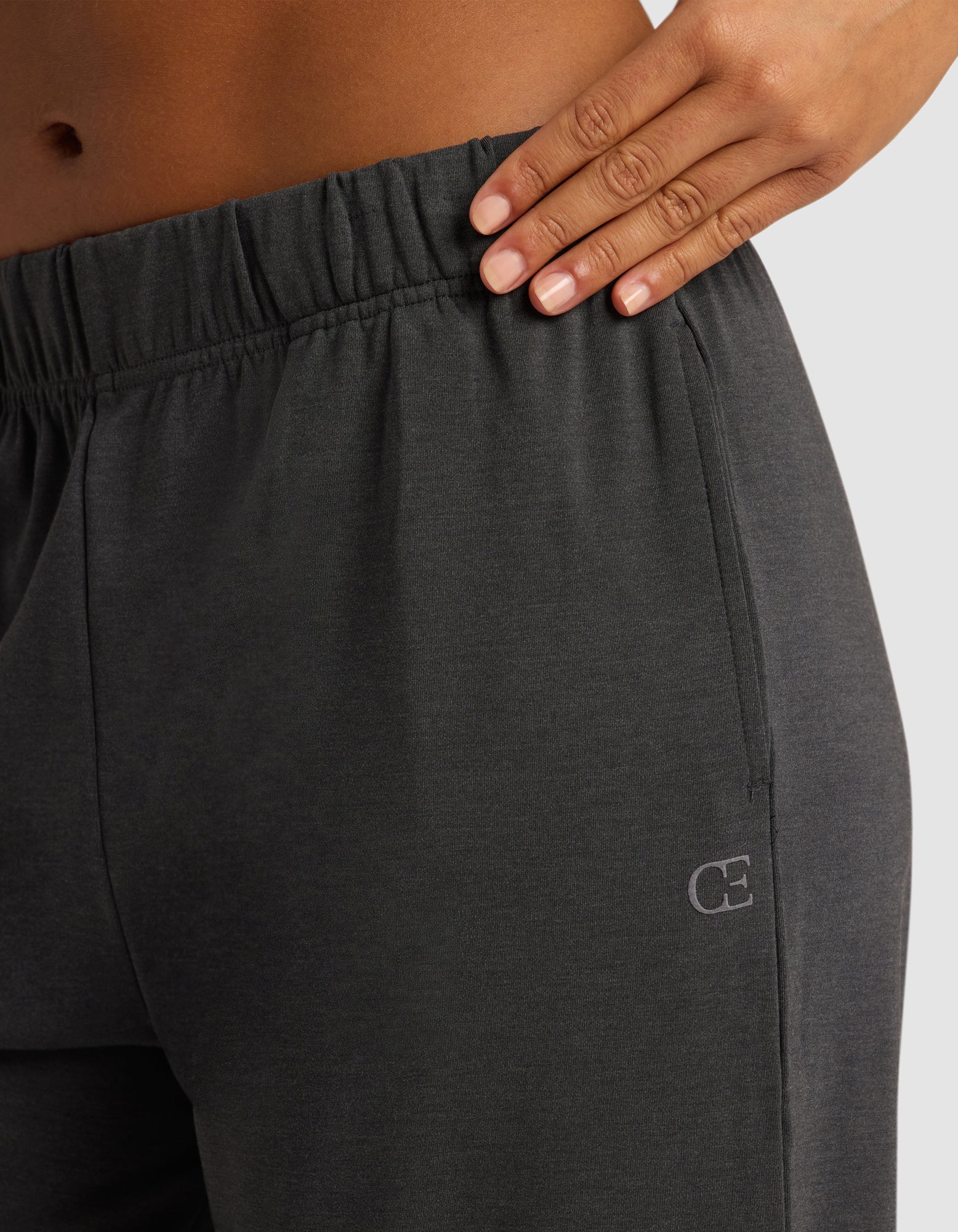 A close-up of a hand resting on the waistband of Cozy Earth's Women's Brushed Bamboo Wide Leg Pull-On Pant in dark gray, featuring an elastic band and a small "CE" logo on the hip against a plain light-colored background.|Color:Charcoal