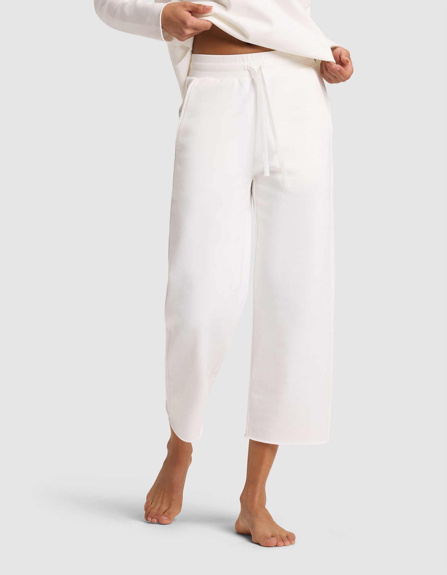 A woman, barefoot on a light background, models Cozy Earth's Women's Chelsea Cropped Pant—loose-fitting, high-waisted, white lounge pants with a drawstring and pockets. Only her lower body is shown. 