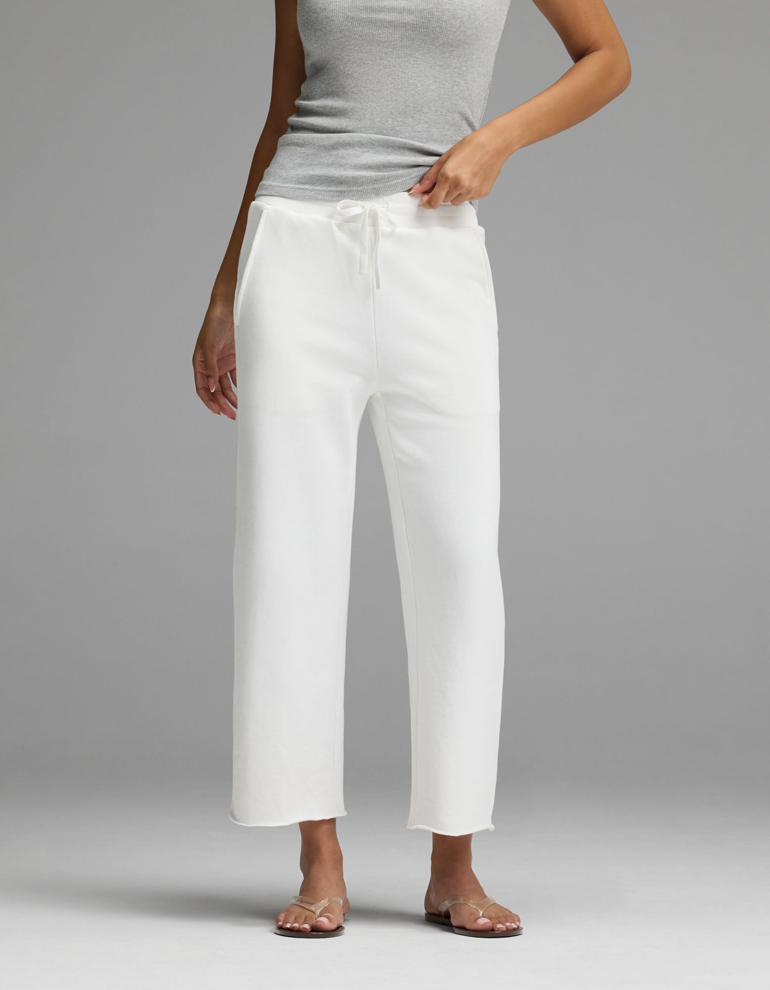 A person wearing the Cozy Earth Women’s Chelsea Cropped Pant in Bone stands on a gray background, with one hand adjusting the waistband. Their face is not visible.