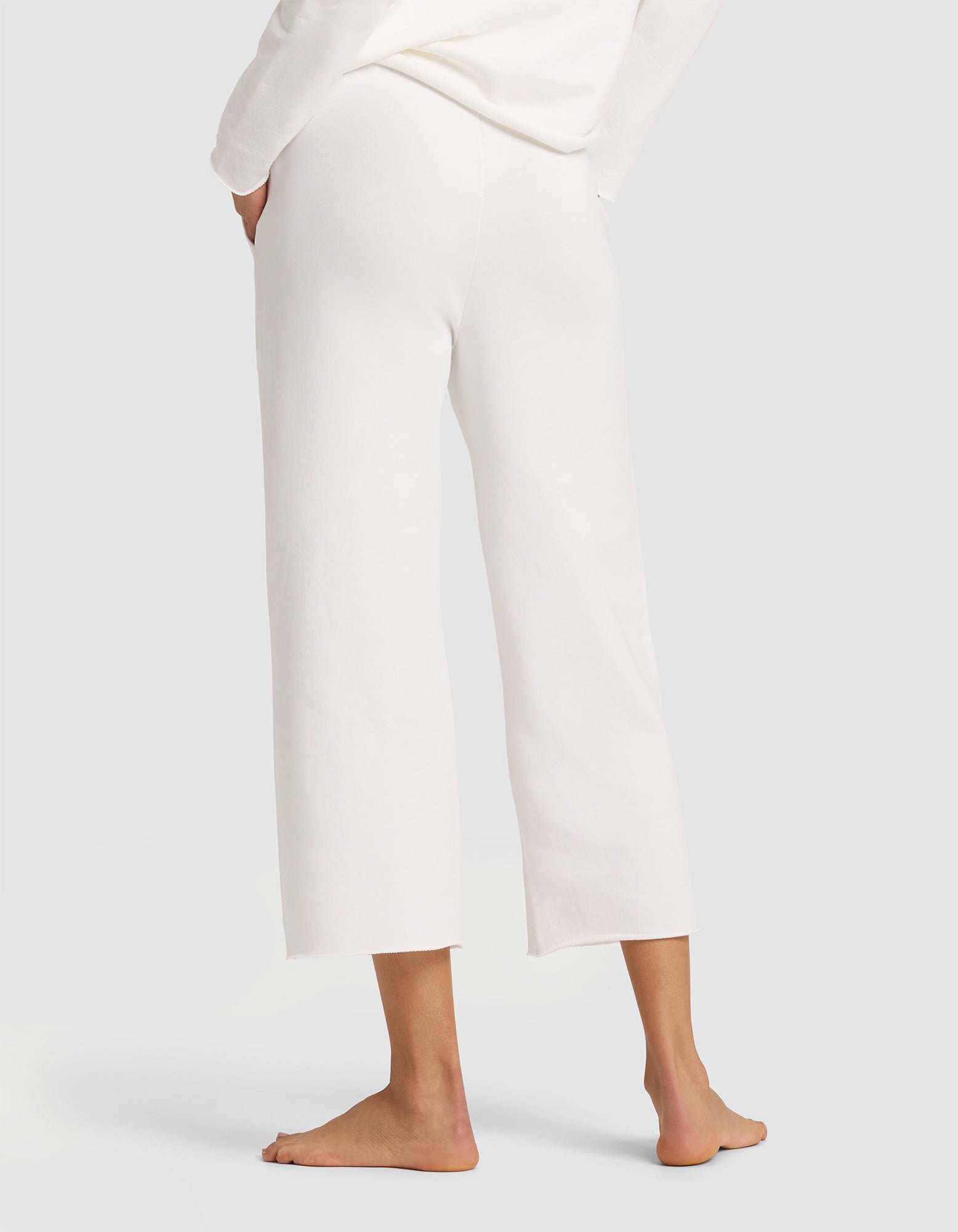 A woman stands barefoot on a light background with her back turned and hands in her pockets, wearing Cozy Earth's Women's Chelsea Cropped Pant and a matching long-sleeve top. 