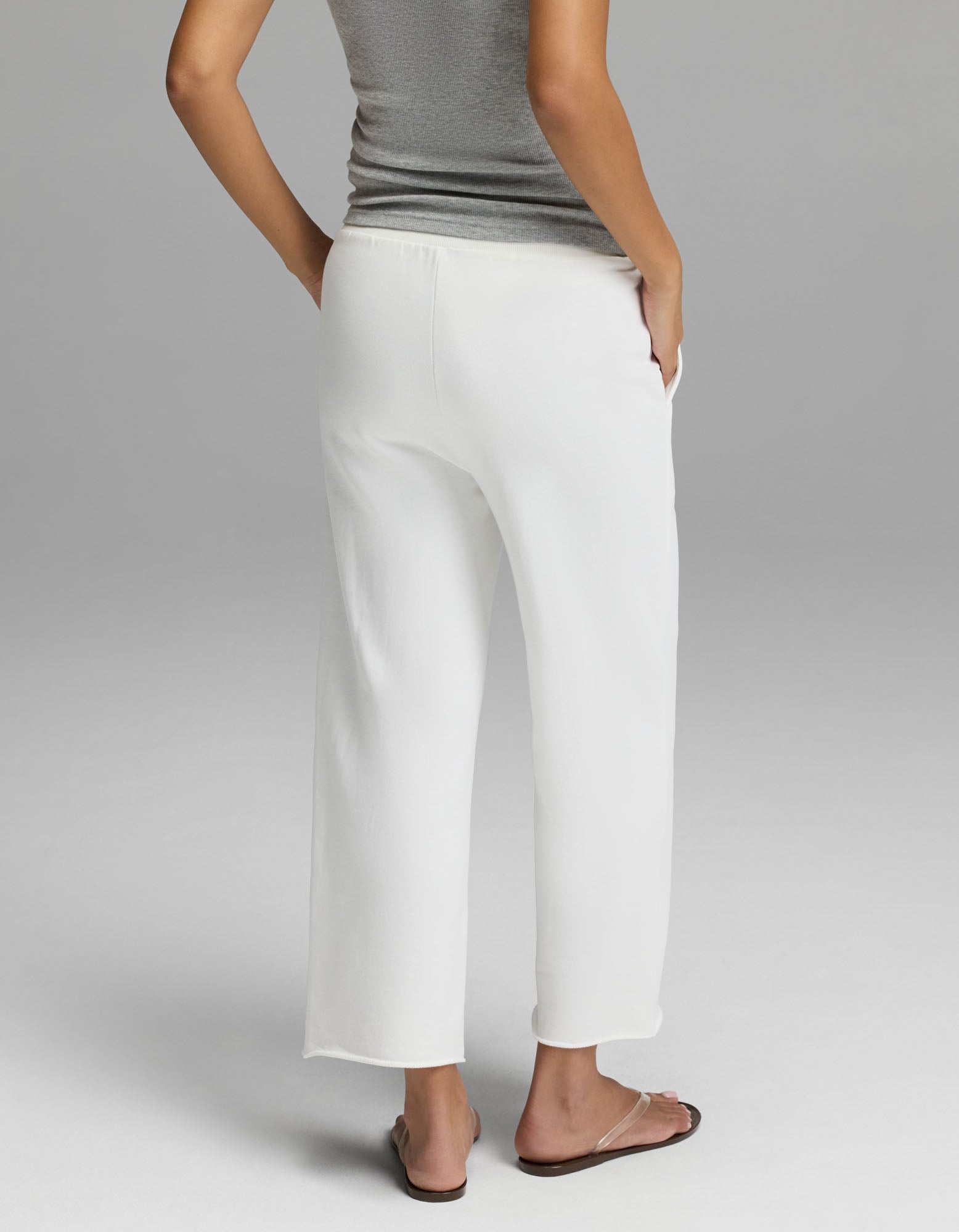 A woman is photographed from behind against a plain light gray background, standing with one hand in the pocket of her loose white Cozy Earth Women’s Chelsea Cropped Pant, paired with a gray sleeveless top and flip-flops.
