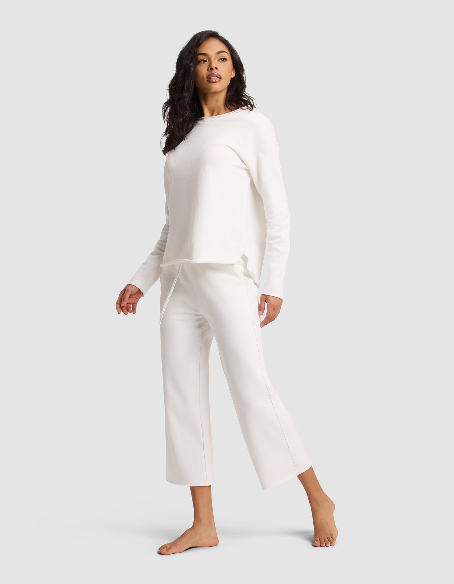 A woman with long dark hair stands barefoot, wearing Cozy Earth's Women's Chelsea Cropped Pant and a matching loose white top, posing against a plain light gray background. 