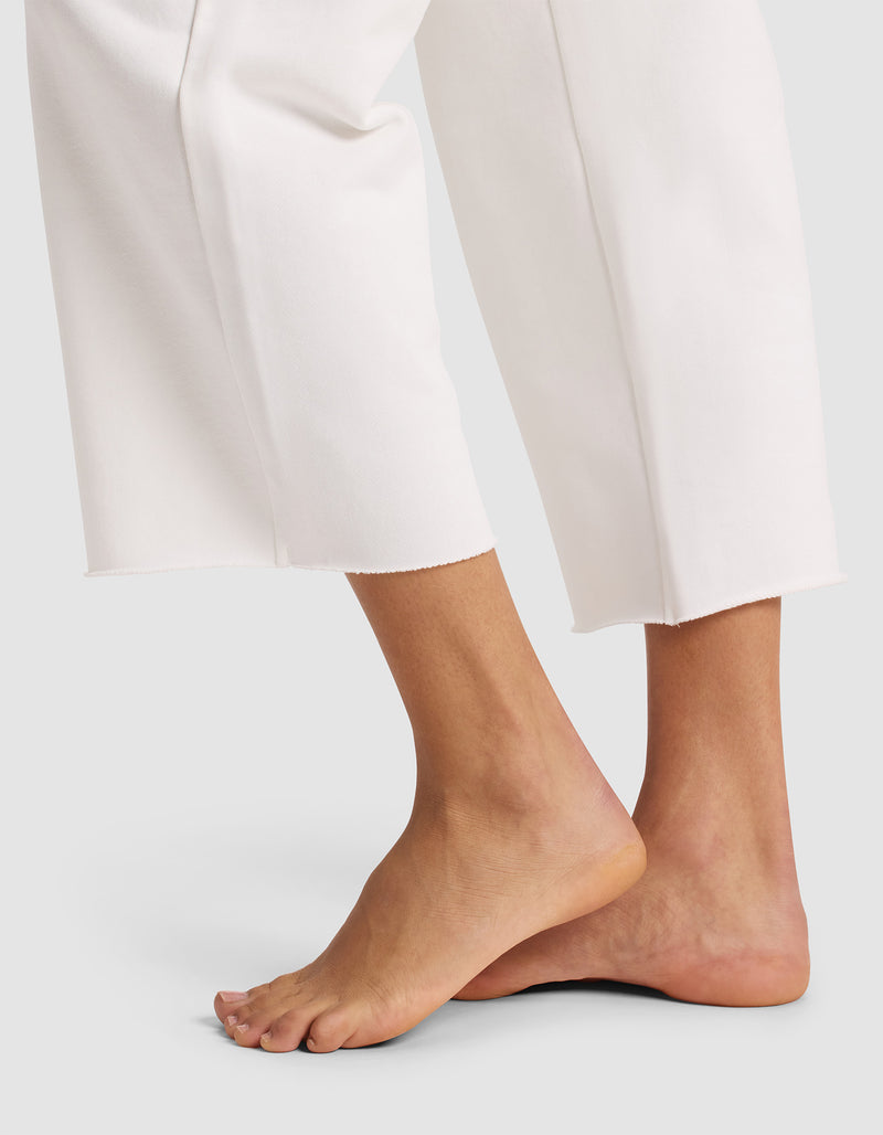 A person stands barefoot on tiptoe against a light background, wearing Cozy Earth Women's Chelsea Cropped Pant—loose-fitting white pants that end above the ankles.