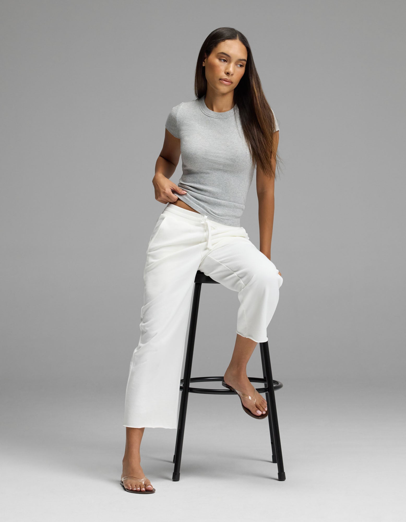 A woman with long brown hair, wearing a light gray t-shirt and the Cozy Earth Women’s Chelsea Cropped Pant in white, sits barefoot on a black stool against a plain gray backdrop.