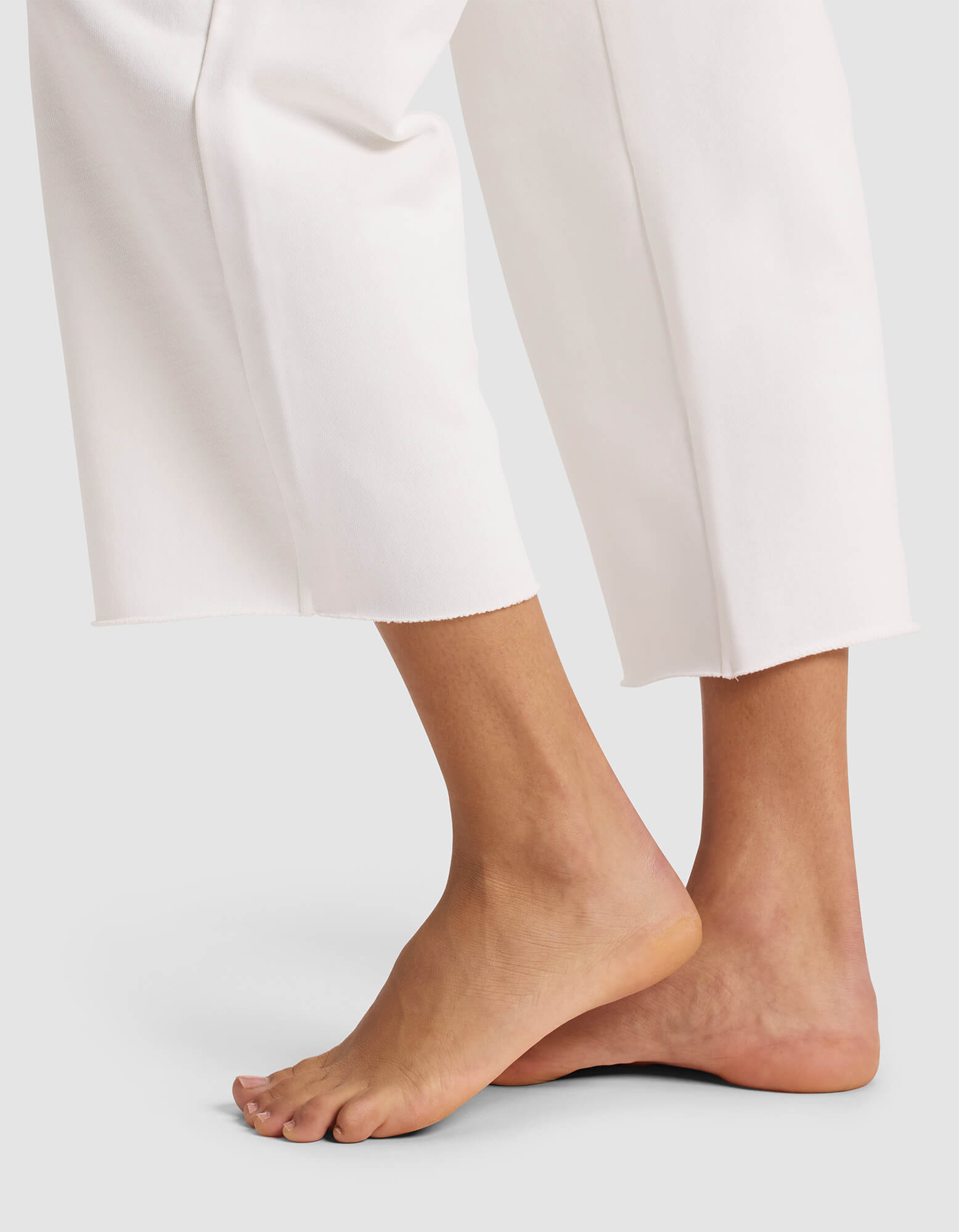 A person stands barefoot on tiptoe against a light background, wearing Cozy Earth Women's Chelsea Cropped Pant—loose-fitting white pants that end above the ankles. 