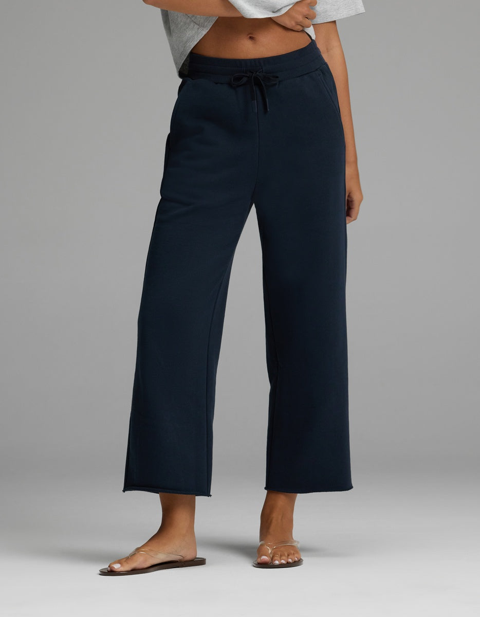 A person is wearing the Cozy Earth Women’s Chelsea Cropped Pant in Eclipse, featuring a drawstring waist and wide-leg silhouette, paired with a light gray cropped top and brown sandals. Only the lower torso and legs are visible, standing against a plain gray background.