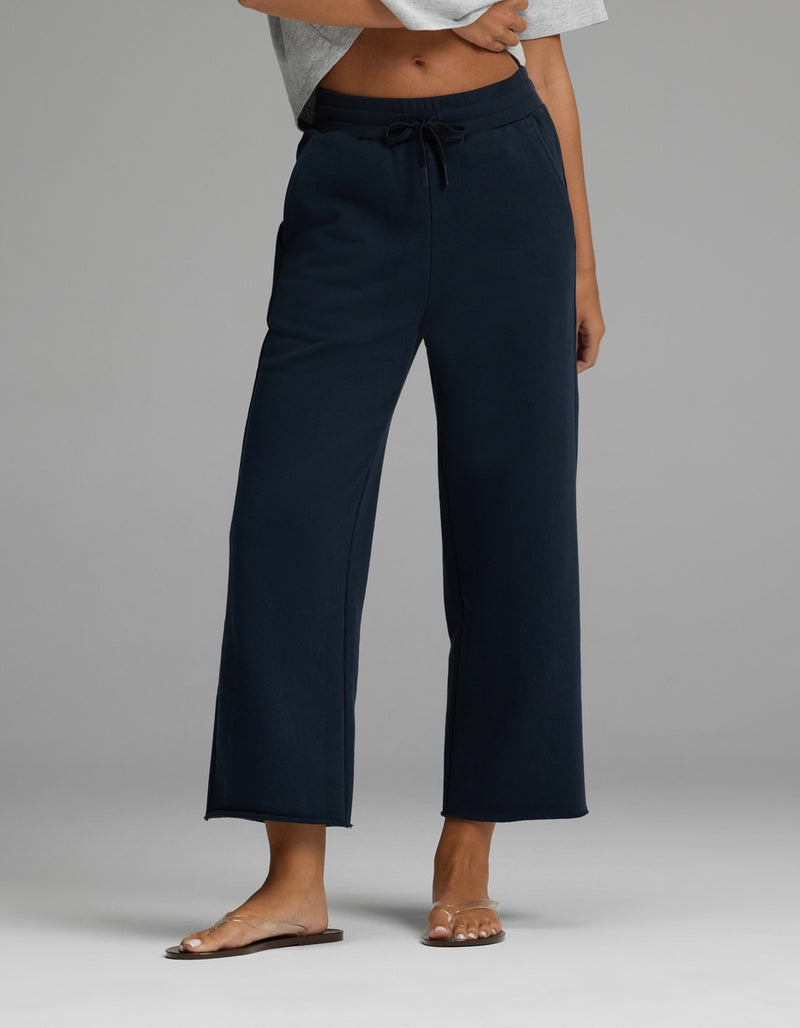 A person is wearing the Cozy Earth Women’s Chelsea Cropped Pant in Eclipse, featuring a drawstring waist and wide-leg silhouette, paired with a light gray cropped top and brown sandals. Only the lower torso and legs are visible, standing against a plain gray background.