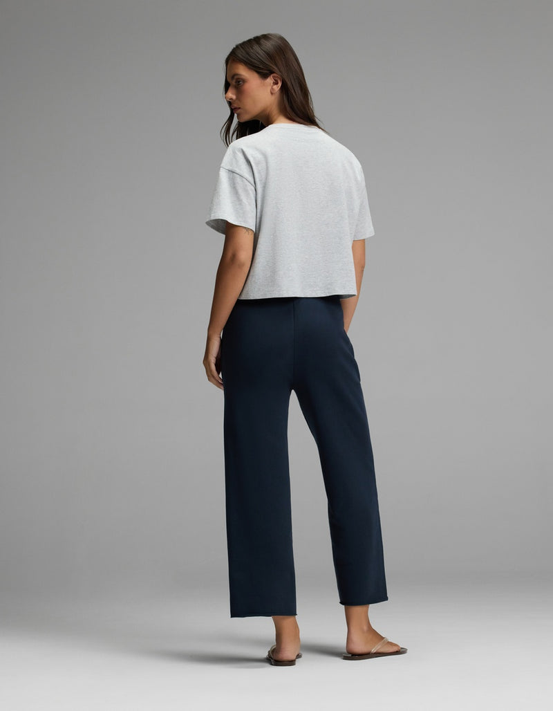 A woman stands facing away against a plain gray background, wearing Cozy Earth’s Women’s Chelsea Cropped Pant in dark blue, paired with a light gray short-sleeve top and brown sandals.