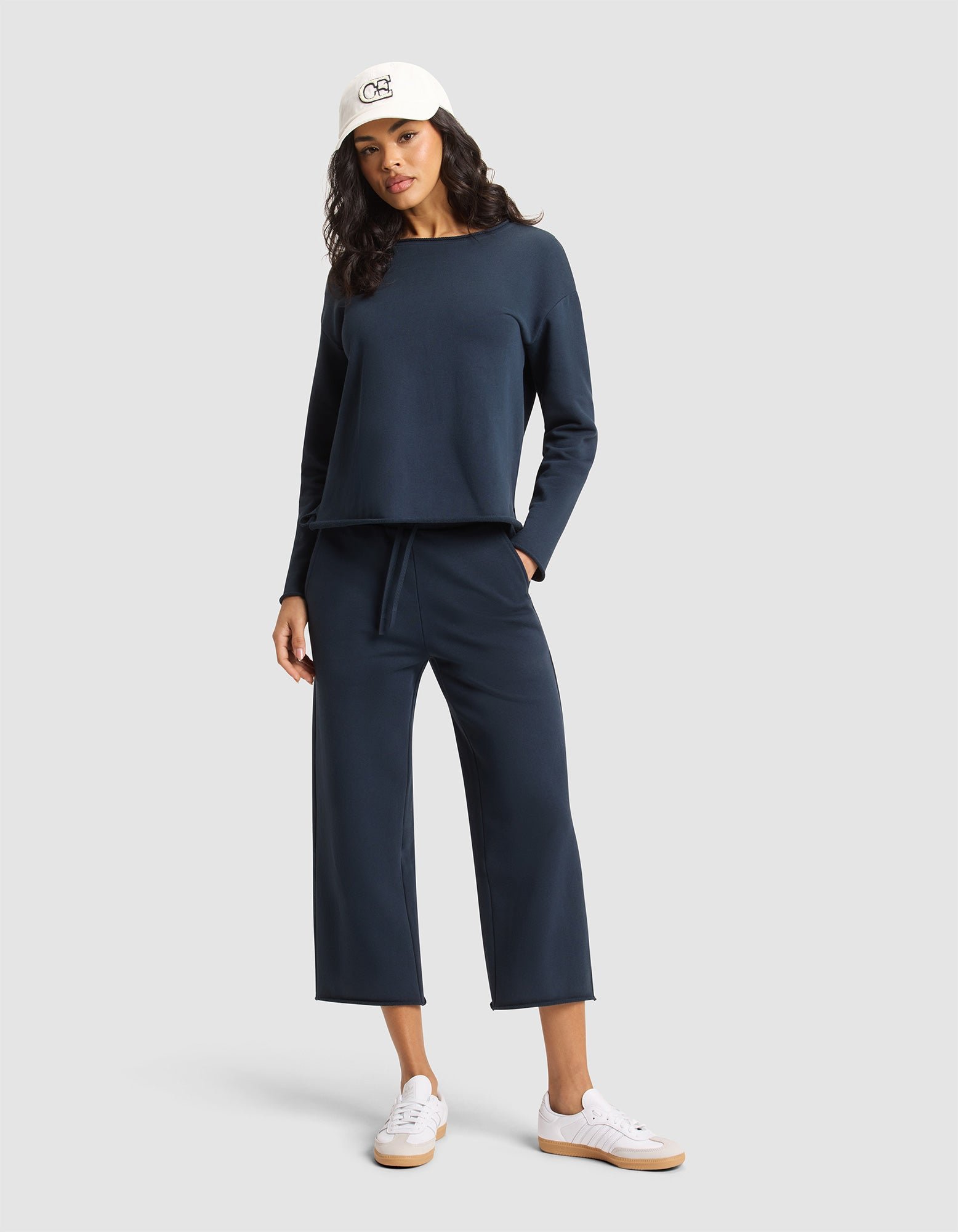 A woman in a white cap, navy long-sleeve top, Cozy Earth Women's Chelsea Cropped Pant, and white sneakers poses with one hand in her pocket against a plain light gray background. |Color:Eclipse