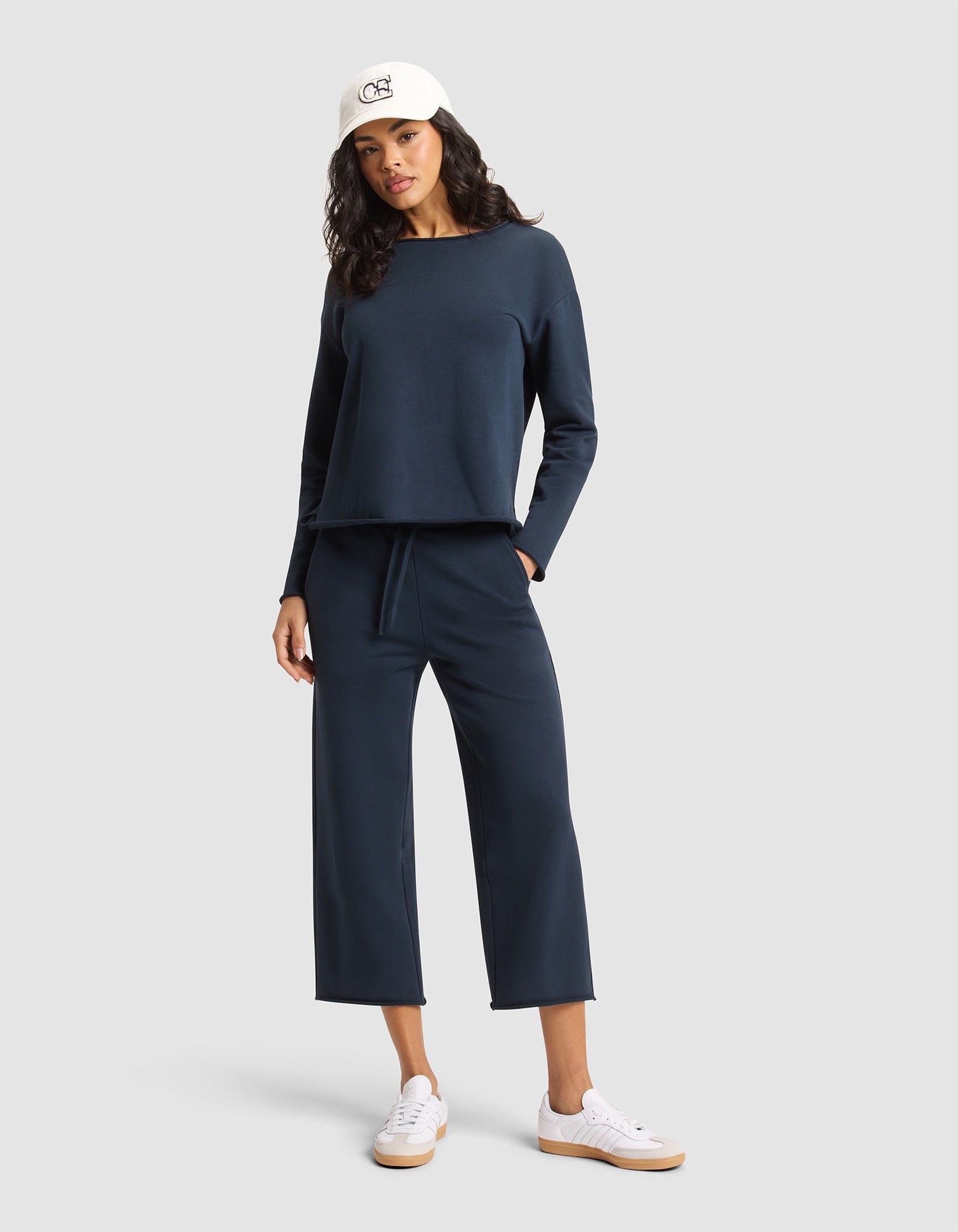 A woman in a white cap, navy long-sleeve top, Cozy Earth Women's Chelsea Cropped Pant, and white sneakers poses with one hand in her pocket against a plain light gray background. 