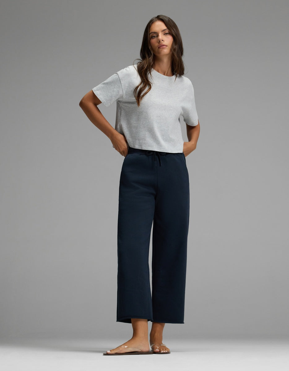 A woman stands barefoot on a gray floor, wearing a light gray short-sleeve t-shirt and Cozy Earth’s Women’s Chelsea Cropped Pant in black, with her hands in her pockets as she looks forward against a plain background.