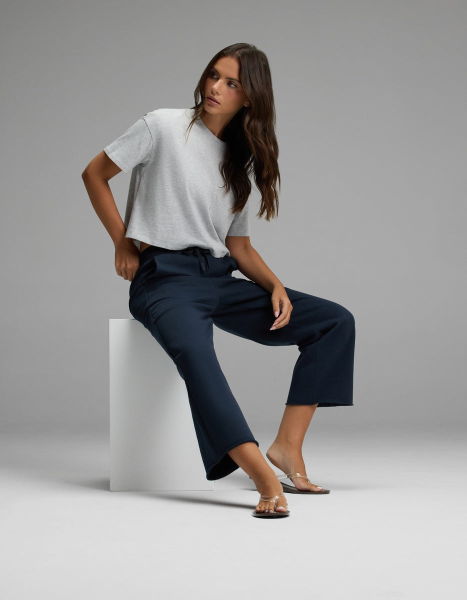 A woman wearing Cozy Earth’s Women’s Chelsea Cropped Pant in navy and a gray t-shirt sits on a white block against a plain gray background, looking to the side with one hand on her hip and sandals on her feet.