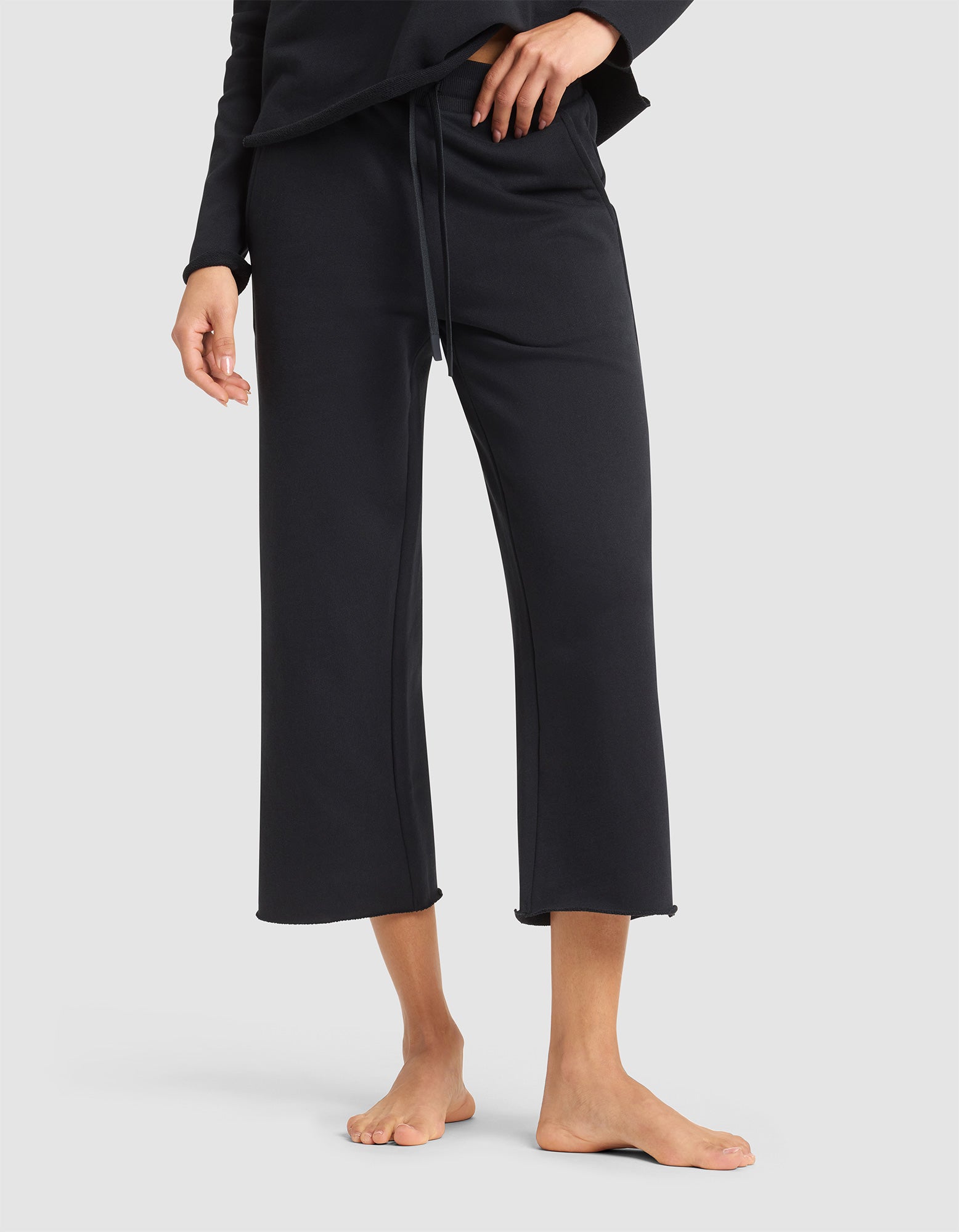 A person is shown barefoot against a plain background, wearing the lower half of Cozy Earth's Women's Chelsea Cropped Pant in black, paired with a matching black top. Only the pants and a portion of the top are visible. 