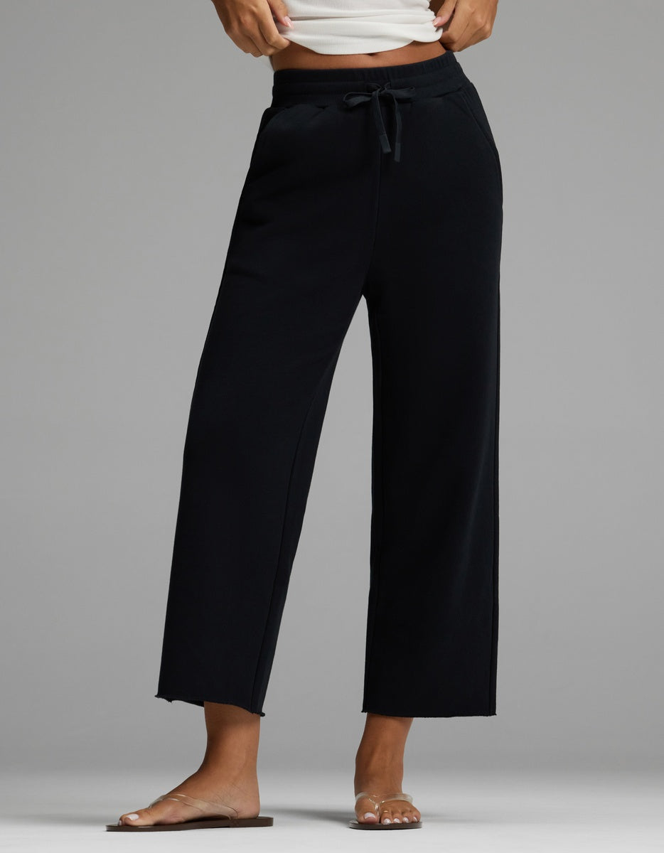 A person is wearing the Cozy Earth Women’s Chelsea Cropped Pant in Jet Black, styled with a white cropped tank top and clear flat sandals. The image highlights the legs and lower torso against a plain gray background.|Color:Jet Black