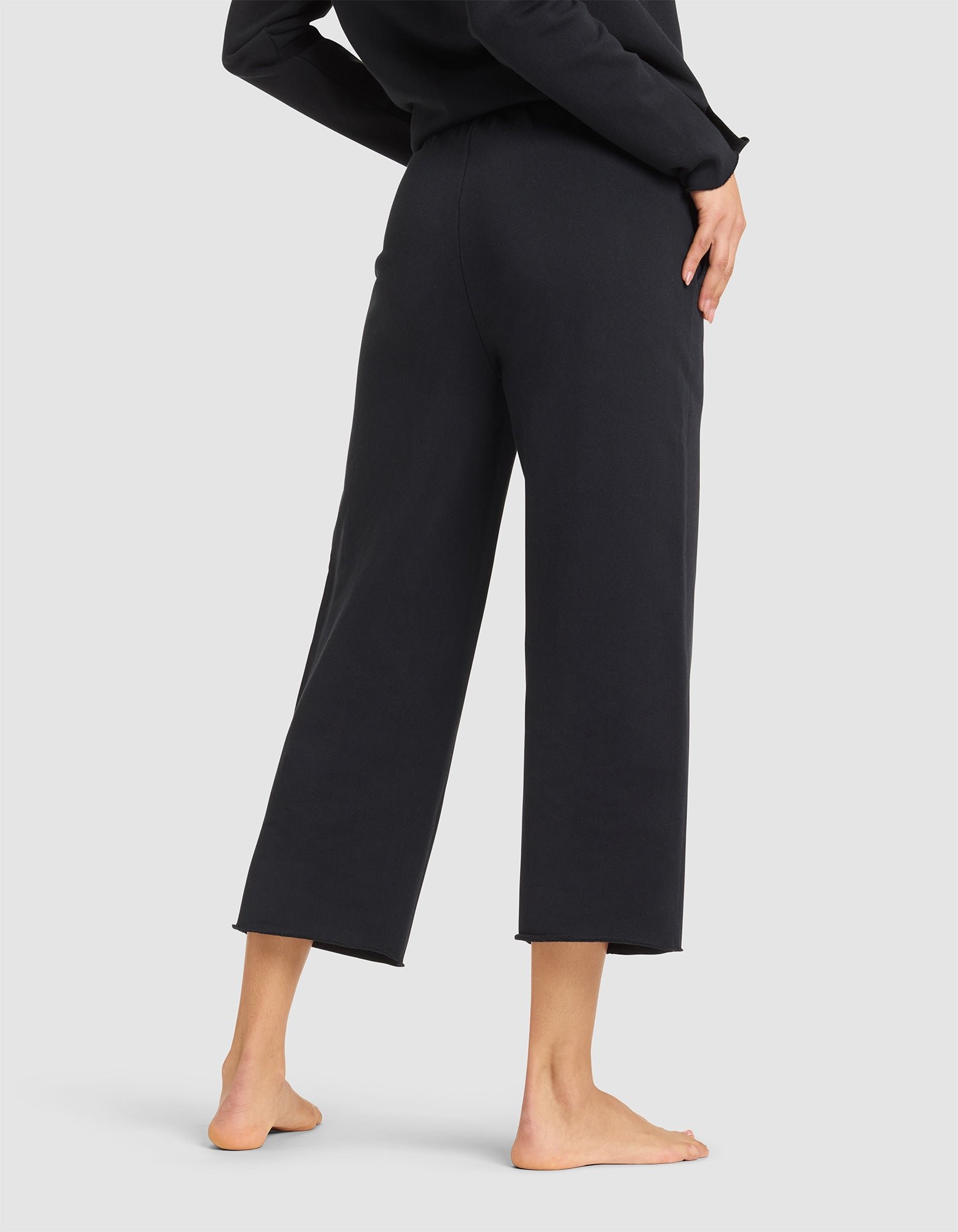 A woman stands barefoot, shown from the waist down, wearing Cozy Earth's Women's Chelsea Cropped Pant in black with a matching top against a plain light background. 