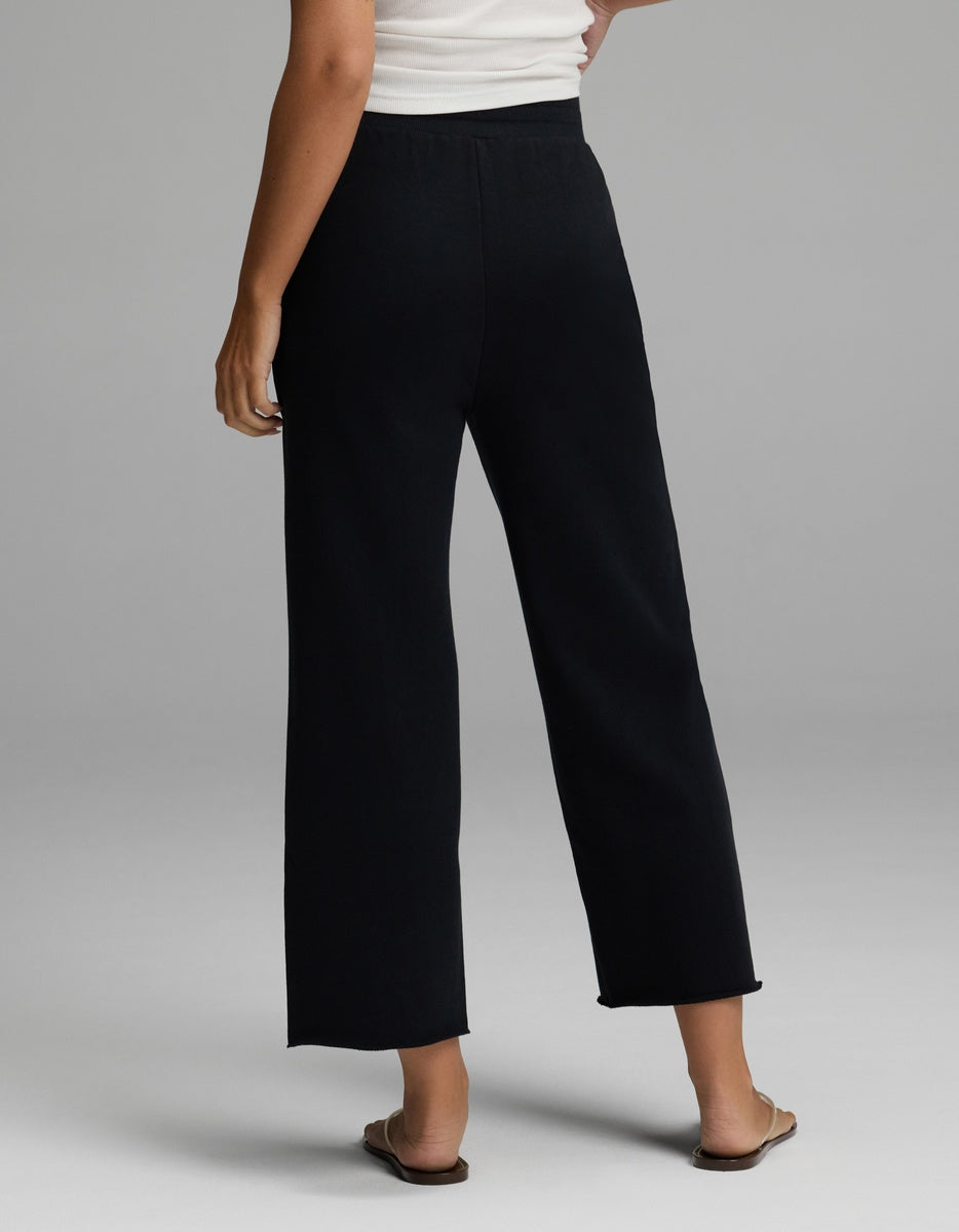 A person is shown from the back standing on a light gray background, wearing Cozy Earth’s Women’s Chelsea Cropped Pant in black, paired with a white sleeveless top and beige flat sandals.