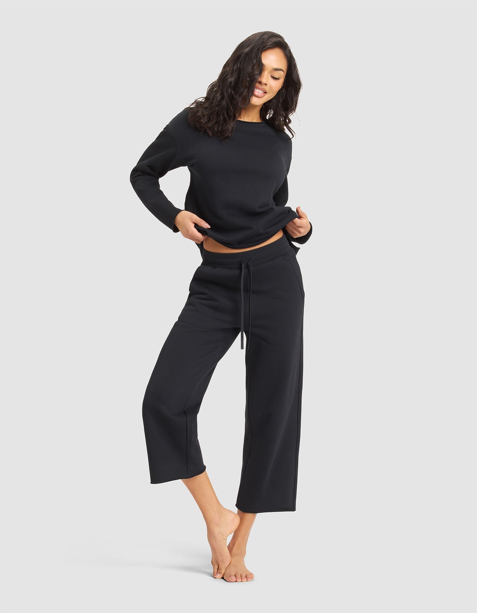 A woman with wavy dark hair wears Cozy Earth's Women's Chelsea Cropped Pant and a loose black top. Barefoot, she stands on one leg, slightly lifts her top, and smiles against a simple light background. 