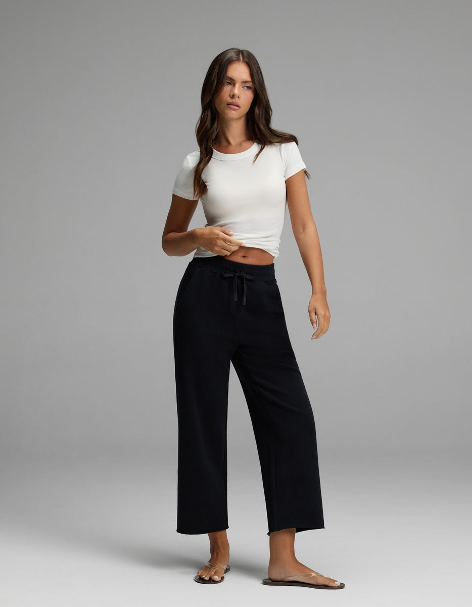 A woman wearing black Cozy Earth Women’s Chelsea Cropped Pants, a white short-sleeve crop top, and sandals stands against a plain gray background.