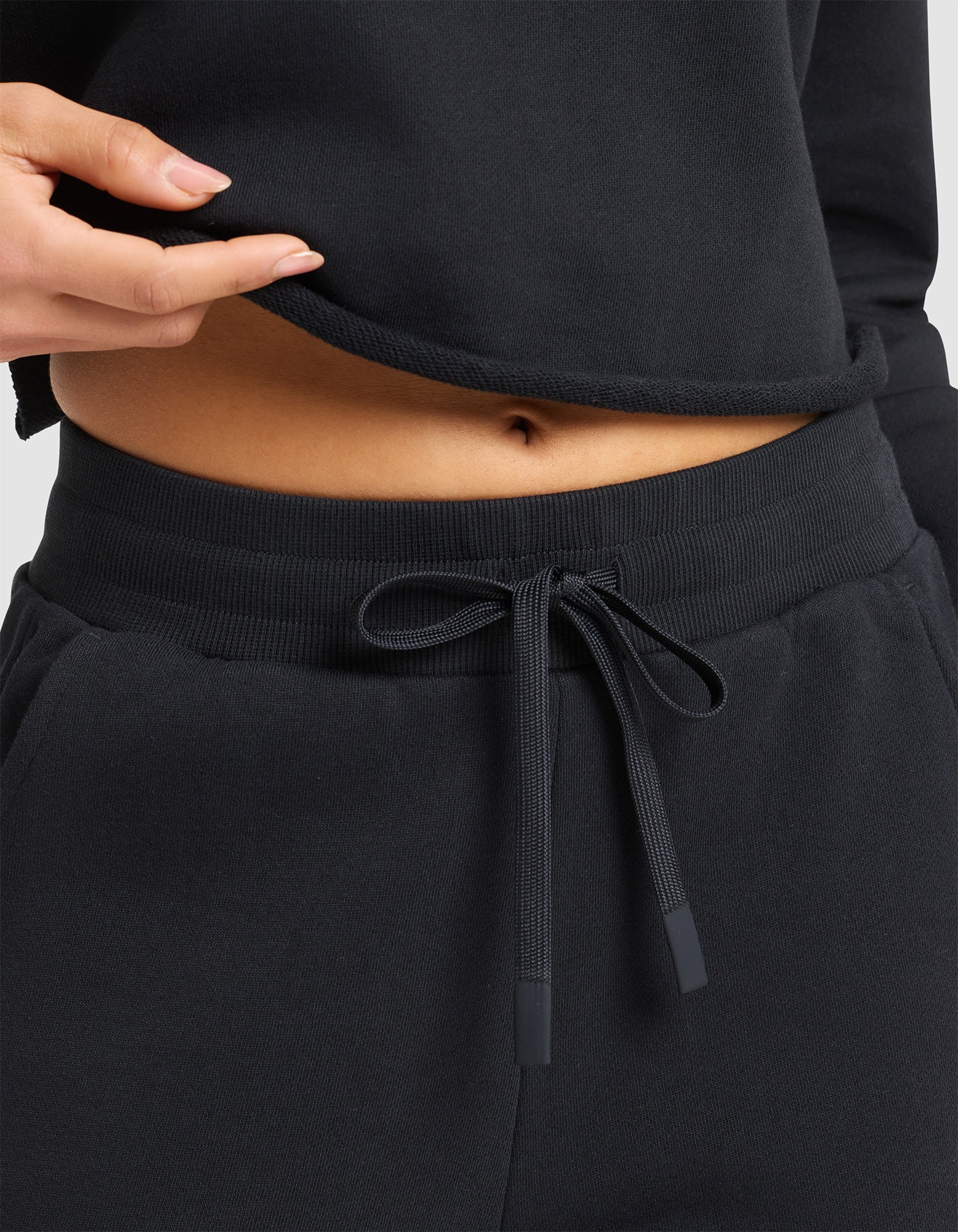 A woman models the Cozy Earth Women's Chelsea Cropped Pant in black, paired with a matching top that's lifted to reveal her bare midriff and navel. 